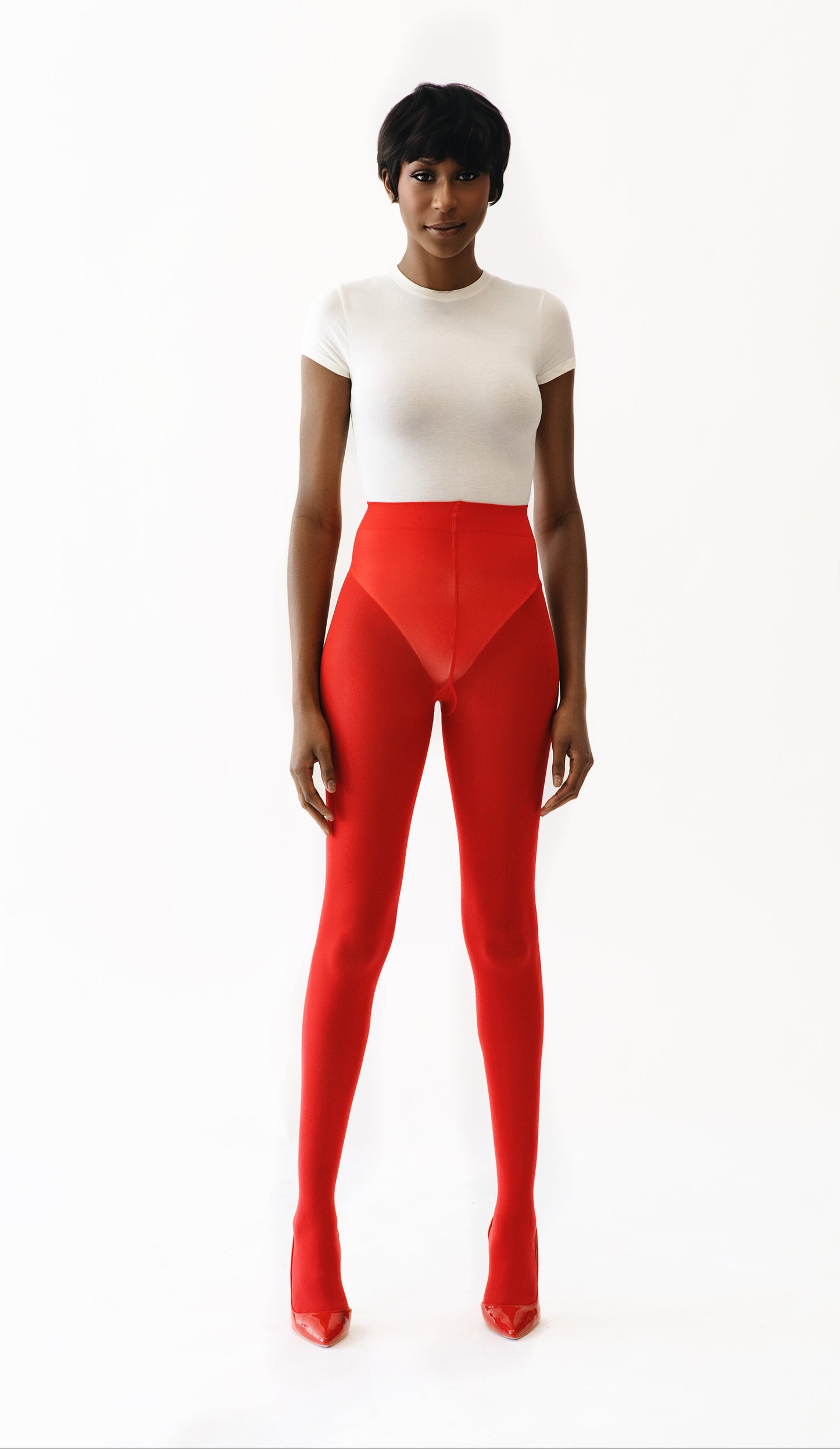 A model stands against a plain white studio background wearing a fitted white short-sleeve top and bright red high-waisted leggings with a geometric front panel.