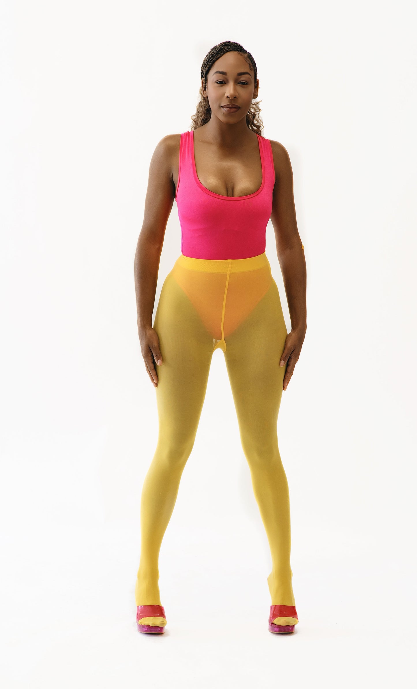 A model stands against a plain white studio background wearing a bright pink bodysuit with yellow tights and pink-and-yellow shoes.