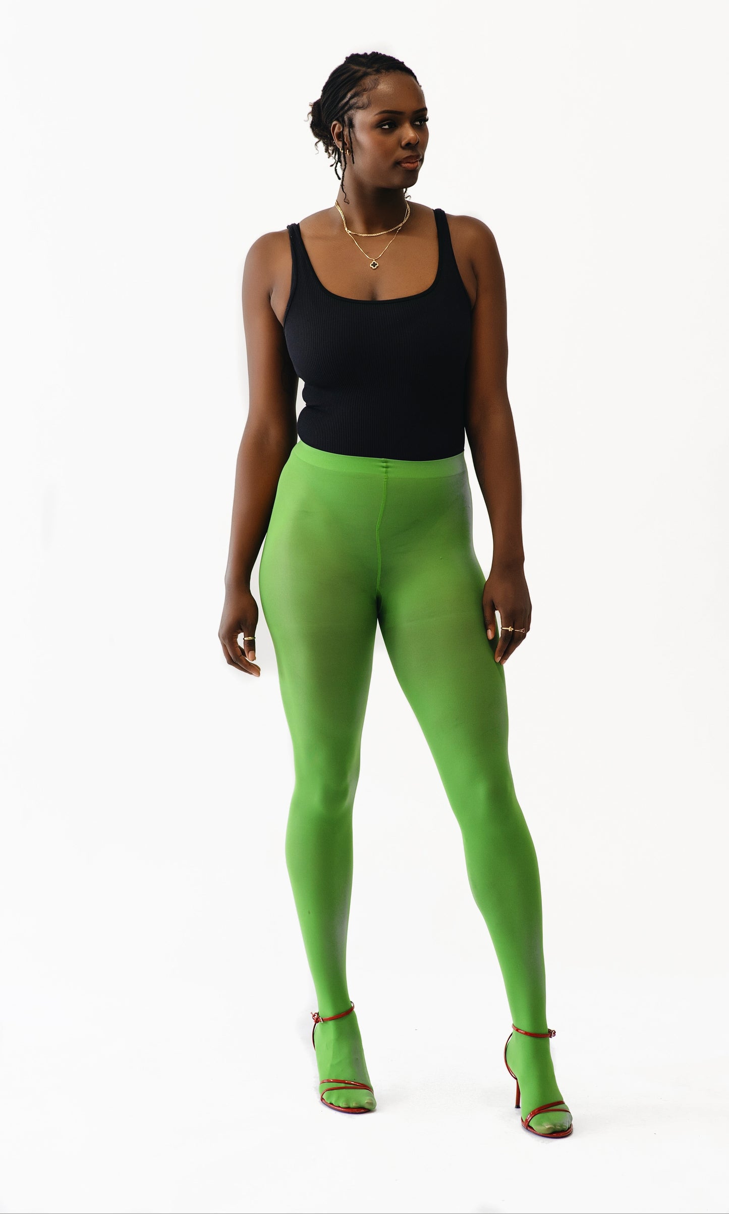 A woman modeling bright lime green full-length leggings with a black tank top and matching green high-heeled sandals, standing against a plain white studio background.