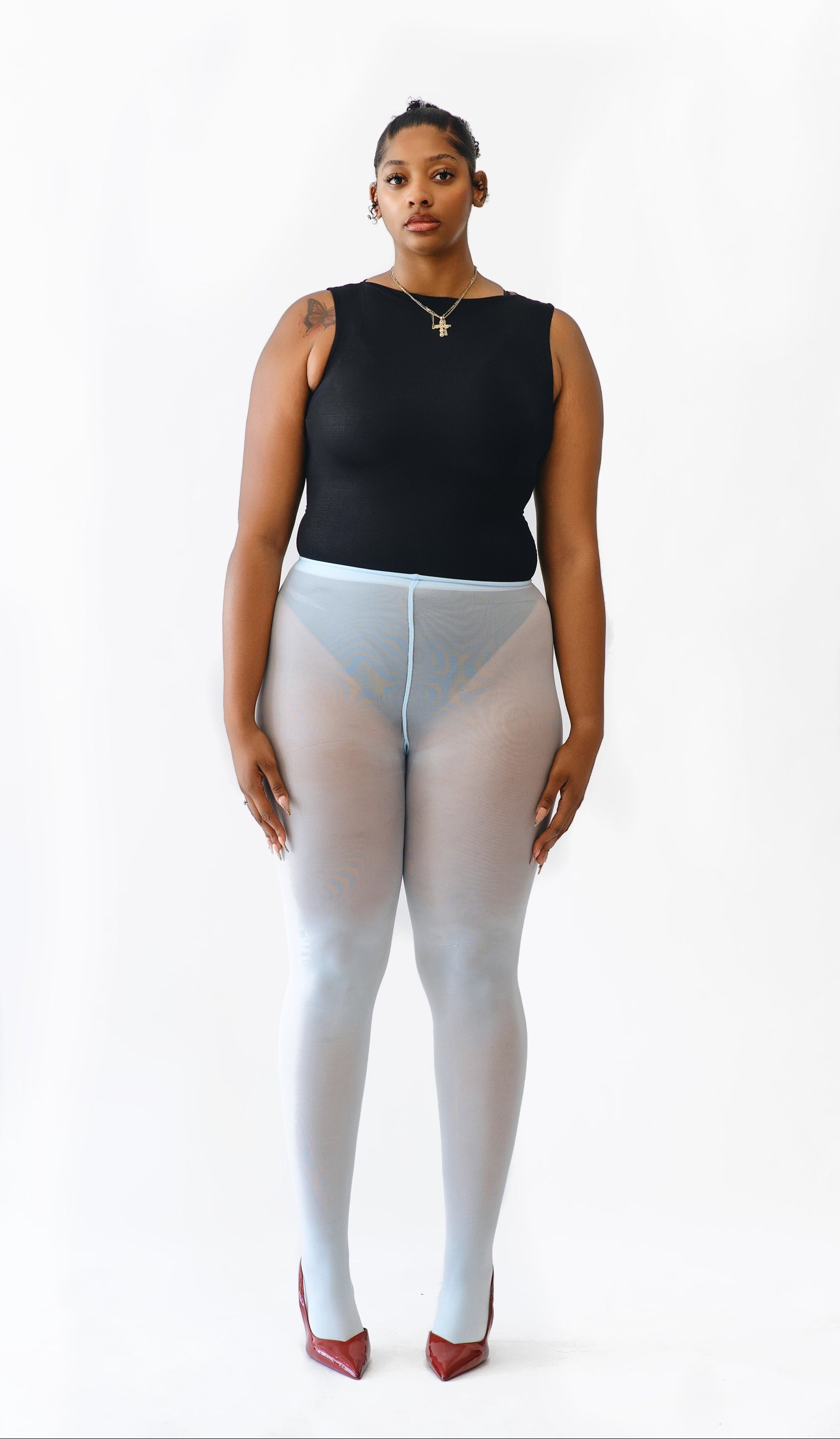 A model stands against a plain white studio background wearing a black sleeveless top, sheer light blue tights, and red pointed heels.