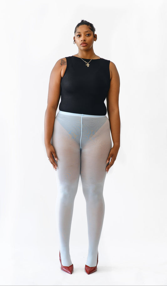 A model stands against a plain white studio background wearing a black sleeveless top, sheer light blue tights, and red pointed heels.